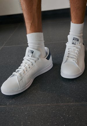 Osoba nosząca białe sneakersy Adidas Stan Smith z granatowym akcentem na pięcie i białe skarpetki, stojąca na ciemnej, kafelkowej podłodze.