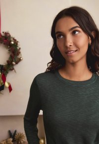 Dark green long-sleeve top with a textured pattern, round neckline, and fitted sleeves. Background features a holiday wreath with decorations.