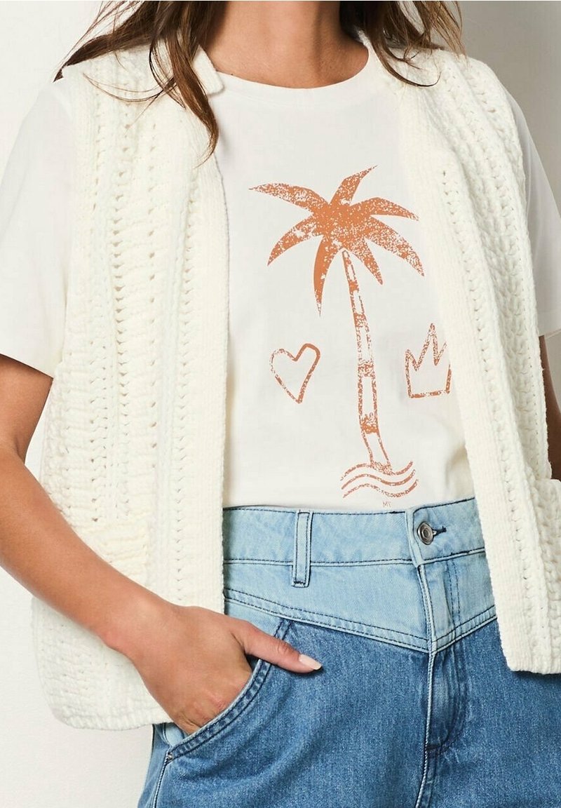 Vit T-shirt med orange palmträd, hjärt- och kronmönster, bärs tillsammans med en krämfärgad stickad väst och tvåfärgade blå högmidjade jeans.