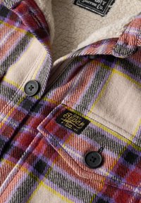 Zbliżenie na czerwoną, fioletową, żółtą i szarą kratowaną kurtkę z czarnymi guzikami i kremową podszewką z polarowego materiału, z metką marki Superdry na kieszeni.