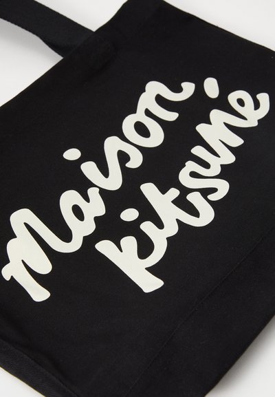 Μαύρη βαμβακερή τσάντα tote με μεγάλα, λευκά, ρέοντα γράμματα που γράφουν "Maison Kitsuné." Απλός σχεδιασμός με ανθεκτικό ύφασμα και μακριά λουριά.