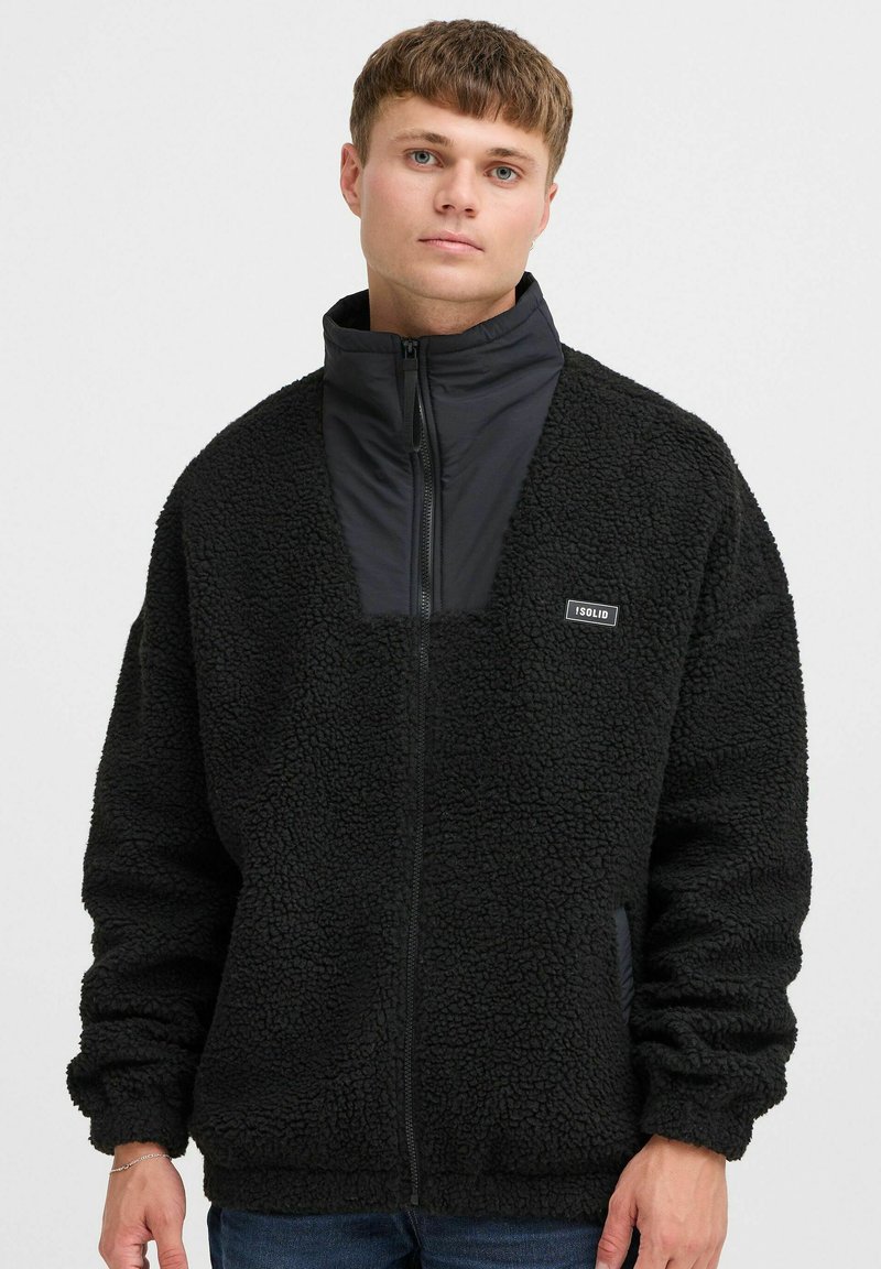 Chaqueta de forro polar negra con cierre frontal, cuello alto y tela suave contrastante en el escote. Presenta una pequeña etiqueta de marca en el pecho.