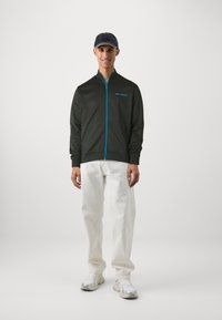Casaco corta-vento verde escuro com detalhes azuis e logótipos, combinado com calças brancas e ténis. Textura suave, corte descontraído e bolsos frontais.