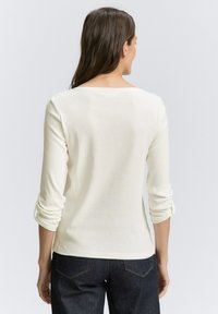 Donna con lunghi capelli scuri che indossa una maglietta bianca a maniche lunghe con maniche arricciate e jeans blu scuro, di spalle rispetto all'obiettivo.