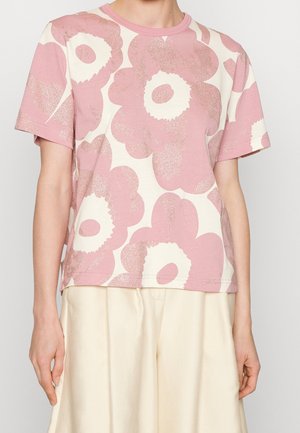 Persona vistiendo una camisa de manga corta con estampado floral en rosa y blanco, y pantalones de color crema, mostrada desde el cuello hasta las caderas.