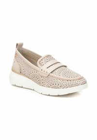 Zapato slip-on de cuero perforado color beige con punta redondeada, adornado con una tira ancha y pequeñas tachuelas, sobre una suela de goma blanca.