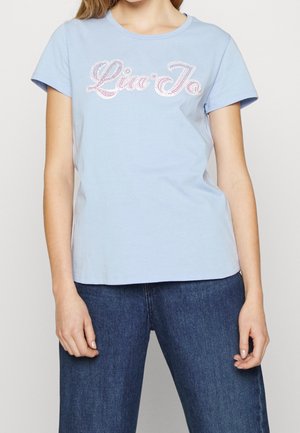 Vrouw in een lichtblauwe t-shirt met korte mouwen, versierd met witte en roze rhinestone tekst, en donkerblauwe spijkerbroek, staand met ontspannen armen.