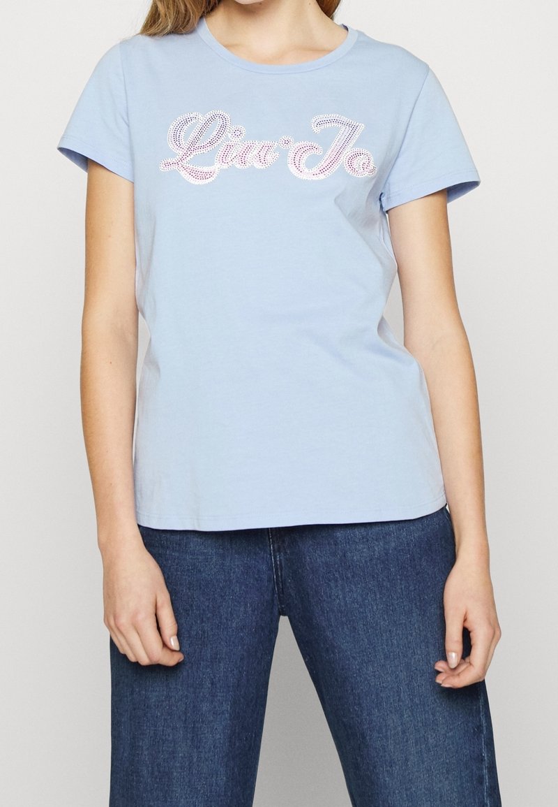 Mujer con una camiseta de manga corta azul claro adornada con texto de strass blanco y rosa, y jeans azul oscuro, de pie con los brazos relajados.