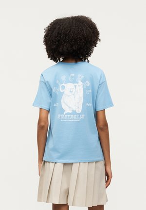 Άτομο με σγουρά μαλλιά που φορά ένα ανοιχτομπλε μπλουζάκι με λευκό κοάλα που κρατάει μια σανίδα του σερφ και μπεζ πλισέ φούστα, με την πλάτη γυρισμένη.