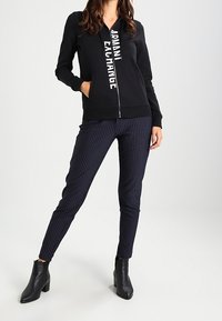 Felpa nera con zip e grafica "ARMANI EXCHANGE" sul davanti, abbinata a pantaloni a righe blu navy e stivali neri alla caviglia.