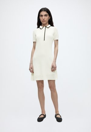 Kvinde med langt mørkt hår bærer en cremefarvet kjole med korte ærmer, knælængde med sort kant ved kraven og sorte Mary Jane-sko.