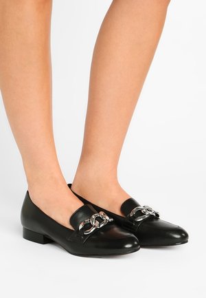 Schwarze Leder-Loafers mit silberner Kettendetailierung, getragen an Zehen und Füßen vor einem schlichten weißen Hintergrund.