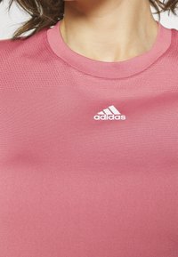 Rosa sporttröja med rund hals, tillverkad av mjukt, elastiskt material. Har en vit Adidas-logga på framsidan. Strukturerad övre del.