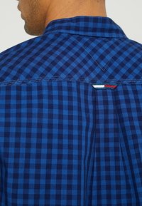 Niebieska koszula w kratę z ciemnoniebieskim i jaśniejszym niebieskim wzorem gingham. Posiada ostre kołnierzyki i kontrastowe detale przeszyć.