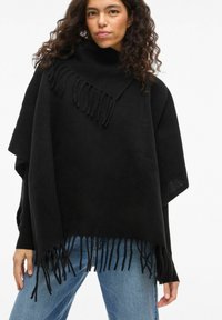 Woman with curly hair wears black fringed poncho over blue jeans against plain white background.