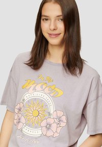 T-shirt en coton violet clair avec un imprimé graphique représentant un soleil et des fleurs, manches courtes et coupe décontractée. Le texte indique "Dans l'Humeur."