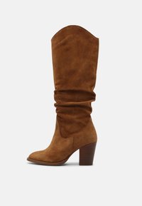 Brązowy zamszowy kozaki za kolano o luźnym fasonie, z ostrym noskiem i obcasem typu słupek. Charakterystyczne dla nich są miękka faktura i zaostrzona górna krawędź.