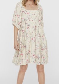 Žena v krémových šatech ke kolen s volánkovými rukávy a růžovo-modrým květinovým vzorem na jednobarevném pozadí.