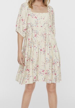 Žena v krémových šatech ke kolen s volánkovými rukávy a růžovo-modrým květinovým vzorem na jednobarevném pozadí.