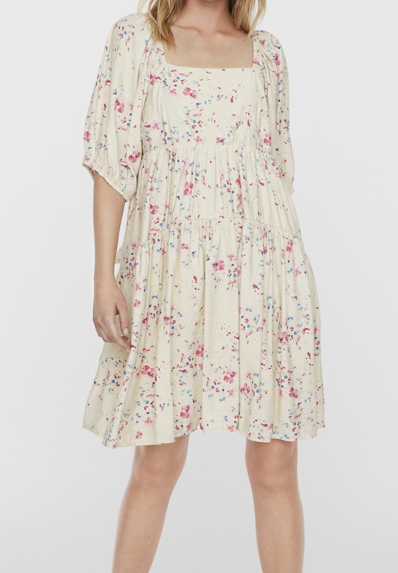 Žena v krémových šatech ke kolen s volánkovými rukávy a růžovo-modrým květinovým vzorem na jednobarevném pozadí.