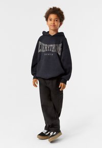 Mustapaitainen huppari, jossa on painettu teksti, yhdistettynä tummaan housuun ja mustiin lenkkikenkiin, joissa on valkoisia yksityiskohtia. Rentoa istuvuutta, pehmeä kangas.