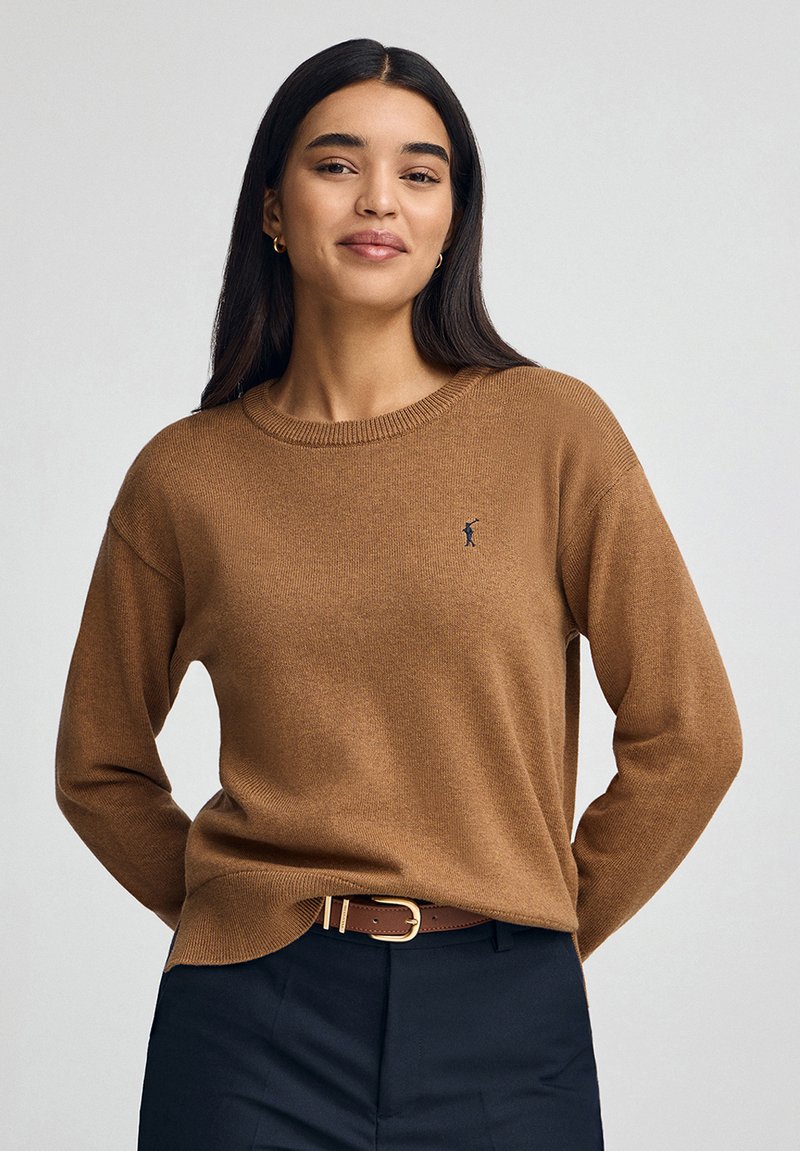 Kobieta o długich ciemnych włosach, ubrana w brązowy sweter z dzianiny i ciemne spodnie, stojąca z rękami za plecami na jednolitym tle.