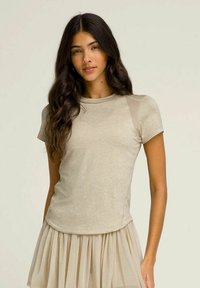 Top beige a maniche corte con scollatura rotonda e pannelli trasparenti sulle spalle, abbinato a una gonna lunga plissettata di un beige chiaro e fluente.