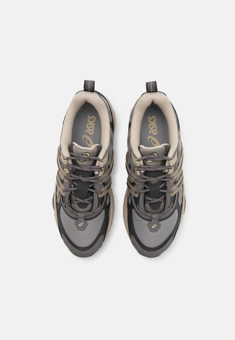 Graue und beige Sportschuhe mit einem Obermaterial aus Mesh, gepolsterter Sohle und dekorativen Schnürsenkeln. Logo auf der Zunge und der Seite sichtbar.