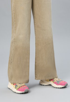 Pantalon ample beige porté avec des baskets colorées arborant des motifs roses, jaunes et en zigzag sur un fond gris.