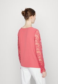 Top cor-de-rosa com mangas longas em renda, apresentando um corpo em tecido suave e detalhes intrincados em renda nas mangas. Design nas costas liso, com decote redondo.