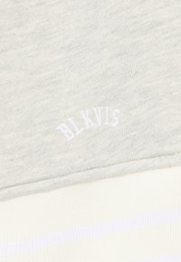 Hellgrauer Stoff mit texturierter Oberfläche, auf dem "BLKVLS" in Weiß gestickt ist. Darunter befindet sich ein gestreiftes Muster in Weiß und blassem Gelb.