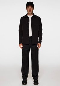 Svart skjorta med knappar framtill och två bröstfickor, bärs över en vit t-shirt med rund hals och svarta byxor, kompletterat med svart keps och skor.