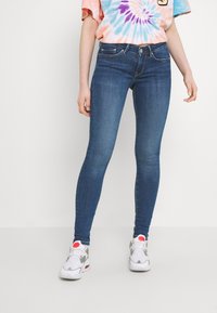 Jeans de mezclilla azul ajustados con una textura suave, que presentan un diseño clásico de cinco bolsillos y una cinturilla estándar. Combinados con zapatillas blancas.