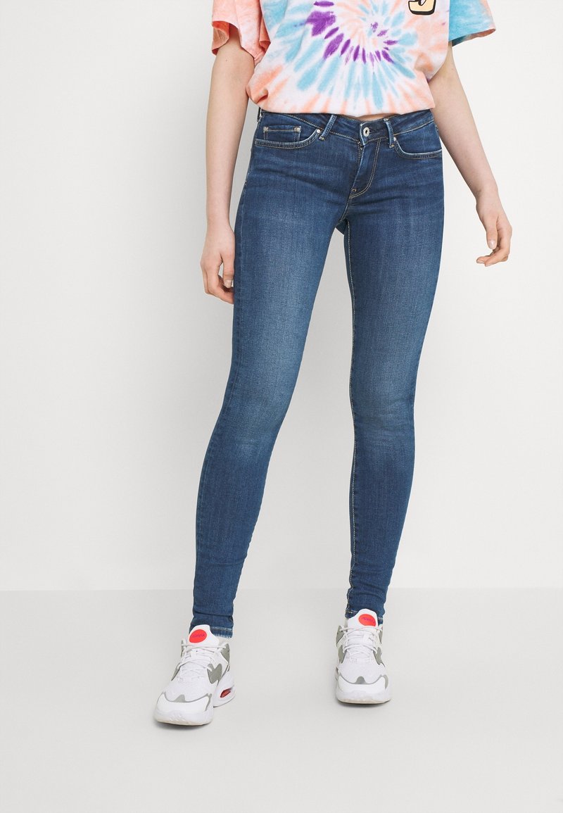 Jeans de mezclilla azul ajustados con una textura suave, que presentan un diseño clásico de cinco bolsillos y una cinturilla estándar. Combinados con zapatillas blancas.