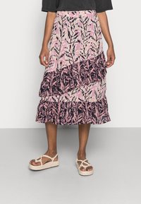Mujer con una falda midi de patrón floral en rosa y negro, con volantes capas y sandalias de cuerda beige, de pie contra una pared blanca.
