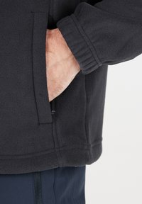 Sort fleecejakke med en blød tekstur, med en lynlåslomme, elastiske manchetter og en lige kant, vist på en models arm.