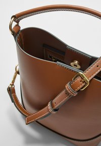 Sac seau en cuir marron avec une sangle cousue et une boucle en métal doré. Il présente un sommet arrondi et une fermeture magnétique pour un accès facile.