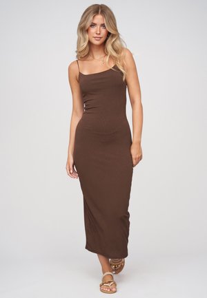 Blonde vrouw die een aansluitende bruine mouwloze midi-jurk en beige sandalen draagt, staand tegen een effen lichte achtergrond.