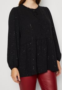 Haut noir à manches longues avec un col volanté, en tissu texturé et avec de subtils accents pailletés. La forme peplum ajoute du volume à l'ourlet.