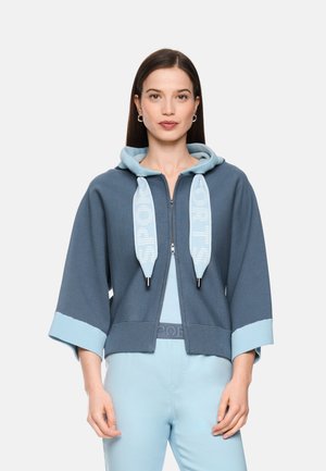 Frau trägt eine blaue kurze Kapuzenjacke mit Reißverschluss, breiten Ärmeln und großen Kordeln, kombiniert mit hellblauen Hosen und einem hellblauen Oberteil.