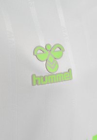Weißer Sportstoff mit einem grünen Hummel-Logo, das ein Bienendesign und fett gedruckten Text zeigt. Glatte Textur mit subtiler Struktur und vertikalen Linien.