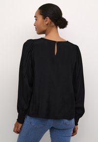 Black blouse with puff sleeves, keyhole back closure, and textured cuffs. Smooth fabric and relaxed fit. Worn with blue jeans.