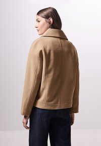 Beige svetr s příměsí vlny s širokým límcem, spuštěnými rameny a prošívanými detaily na zadní straně. Uvolněný střih, v kombinaci s tmavými džíny.
