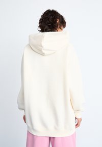 Une personne avec des cheveux courts et crépus de couleur foncée portant un sweat à capuche crème surdimensionné et un pantalon rose, de dos devant un fond blanc uni.
