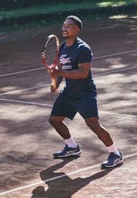 Τενίστας με μπλε πουκάμισο και σορτς, κρατώντας ρακέτα. Φορά μπλε παπούτσια με λευκές λεπτομέρειες και ψηλές κάλτσες μέχρι το γόνατο σε γήινη επιφάνεια.