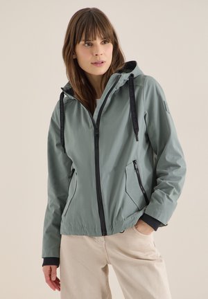 Kobieta ubrana w jasnoszarą kurtkę z kapturem zapinaną na zamek błyskawiczny z czarnymi sznurkami oraz beżowe spodnie, stojąca z jedną ręką w kieszeni.