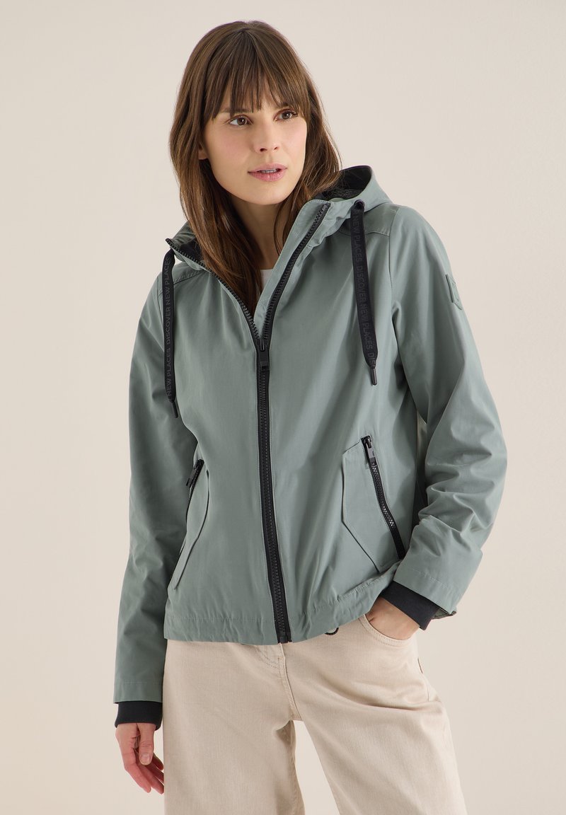 Femme portant une veste à capuche zippée gris clair avec des cordons noirs et un pantalon beige, debout avec une main dans la poche.