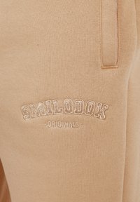 Beige Jogginghose mit weichem Material, elastischem Bund, seitlichen Taschen und aufgesticktem "SMILODOX ORIGINALS" Logo in einer kontrastierenden Farbe.