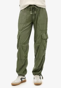 Pantalons cargo en coton vert olive, dotés d'une taille élastique avec cordon de serrage, de plusieurs grandes poches et de jambes fuselées. Une paire de baskets en dessous.