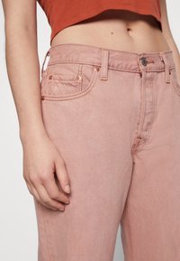 Jean en denim rose avec un design à cinq poches, des garnitures en ton cuivre et une texture lisse. La taille est équipée de passants de ceinture et d'une fermeture à bouton.